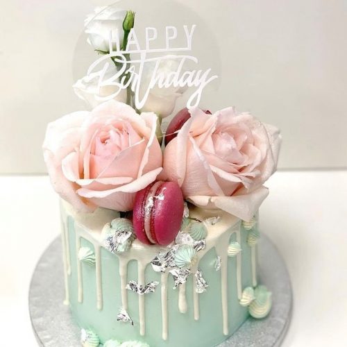 Die Geburtstagstorte hat eine hellblaue Buttercreme sowie einen weißen Drip. Rote Macarons und rosane Rosen gestalten die Torte.